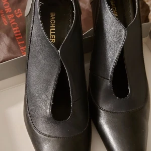 Svarta pumps från Bachiller Madrid - Säljer ett par svarta pumps från Salvador Bachiller Madrid i skinn med unik V-formad öppning framtill. Skorna har spetsig tå och smal klack som ger en elegant siluett. Perfekta för dig som gillar klassisk men ändå modern stil. Svart och grått skinn.