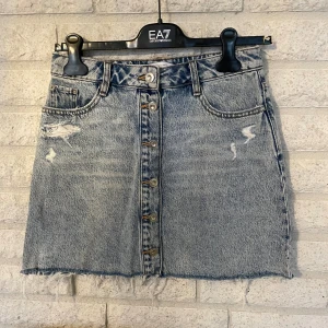 Blå jeanskjol med knappar från Zara - Snygg blå jeanskjol från Zara med slitna detaljer, knappar framtill och rå kant nertill. Klassisk femficksmodell i denim med en cool, avslappnad vibe. Perfekt för dig som gillar en trendig och avslappnad stil.