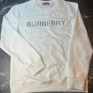Vit Burberry sweatshirt med logga - Säljer en vit sweatshirt från Burberry med klassisk logga och texten 'London England' framtill. Tröjan har långa ärmar, rund halsringning och ribbade muddar. Loggan har Burberrys ikoniska rutiga mönster i bokstäverna. Använt bara en gång