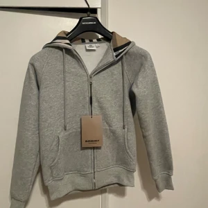 Grå Burberry hoodie  - Säljer en grå hoodie från Burberry i storlek S. Tröjan har dragkedja framtill, snörning vid huvan och klassiskt Burberry-rutigt mönster på insidan av huvan. Mjuk bomullskänsla och ribbade muddar vid ärmslut och nederkant. Perfekt för dig som gillar stilrena detaljer.