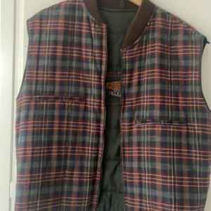 Rutig dunväst från Gant - Säljer en rutig dunväst från Gant med broderad text och flaggor på ryggen. Västen har mörkblå, gröna och röda toner med inslag av gult och brunt. Den är quiltad och har två fickor framtill samt en brun krage. Perfekt för dig som gillar preppy college-stil.