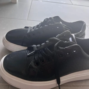 Svarta sneakers från Calvin Klein - Snygga svarta sneakers från Calvin Klein med vit platt sula och rund tå. Skorna har snörning och en blank finish med subtilt mönster på sidorna. Perfekt för dig som gillar stilrena och moderna skor med en touch av designer.