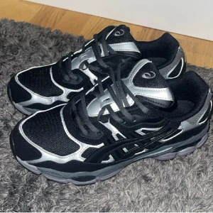 Svarta och silver Asics sneakers - Säljer ett par snygga Asics sneakers i svart och silver med coola detaljer och mesh-ytor. Skorna har rund tå, platt sula och snörning framtill. Perfekt för dig som gillar sportig stil och vill sticka ut med metallic-effekter. Ikonisk Asics-logga på tungan och sidan.