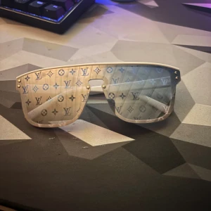 Louis Vuitton vita shield-solglasögon - Säljer ett par vita shield-solglasögon från Louis Vuitton med spegellinser och ikoniskt LV-monogram över hela glaset. Ramen är vit och har en modern, sportig vibe. Perfekta för dig som vill sticka ut med en lyxig och trendig accessoar. Kom med bud på dom 