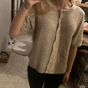 Beige stickad kofta från lager 157, S - Supermysig beige stickad kofta från lager 157 med korta ärmar och pärlknappar framtill. Koftan har en avslappnad passform och ribbade kanter vid ärmslut och nederkant. Perfekt att slänga på sig över en t-shirt eller linne.