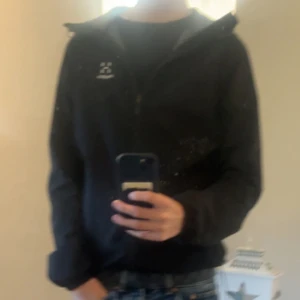 Svart vindjacka från Haglöfs - Svart vindjacka från Haglöfs med dragkedja framtill och hög krage. Jackan har en stilren design med diskret logga på bröstet och långa ärmar. Perfekt för dig som gillar outdoor och vill ha en enkel, funktionell jacka. Dragkedjan e lite sisådär men funkar 