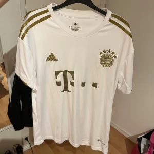 Vit FC Bayern München tröja Adidas S - Säljer en vit FC Bayern München fotbollströja från Adidas i storlek S. Tröjan har guldiga detaljer, klubbmärke och Adidas-logga på bröstet samt tre guldiga ränder på axlarna. Tillverkad i lätt och ventilerande polyester.