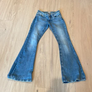 Blå bootcut jeans från Gina Tricot - Bootcut jeans från Gina Tricot i klassisk blå tvätt med slitningar och snygga detaljer på bakfickorna. Modellen har låg midja och breda ben som ger en retro vibe. Tillverkade i jeansmaterial med dragkedja och knapp framtill.