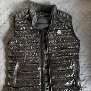 Svart dunväst från Moncler, M - Snygg svart dunväst från Moncler med glansig yta och quiltad design. Västen har två fickor med dragkedja och klassisk Moncler-logga på bröstet. Perfekt att slänga över en hoodie för extra värme. Stängs med dragkedja framtill.