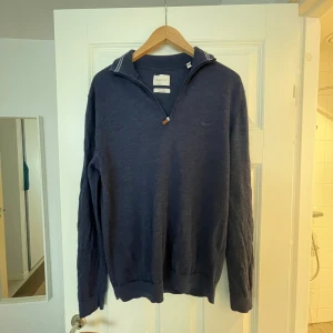 Marinblå merinoulltröja från GANT - Säljer en marinblå långärmad tröja från GANT i extra fin merinoull. Tröjan har en stilren half zip-dragkedja med läderdetalj och diskret broderad logga på bröstet. Perfekt för lager-på-lager och snygg till jeans eller chinos.