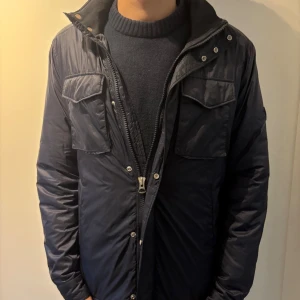 Mörkblå quility jacka från J.Lindeberg - Säljer en mörkblå pufferjacka från J.Lindeberg med quiltad design, två stora bröstfickor med lock, dragkedja och tryckknappar framtill. Jackan har lång ärm och patch med JL-logga på armen. Fodret är i syntet och jackan passar perfekt för kyliga dagar.