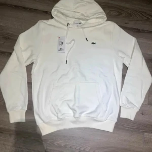 Vit Lacoste Hoodie - Säljer denna stilrena, nya och riktigt snygga vita Lacoste hoodien, passar perfekt till allt, dock är den liten i storleken, står XL men passar (M-L), skriv gärna vid intresse och för mer information! 