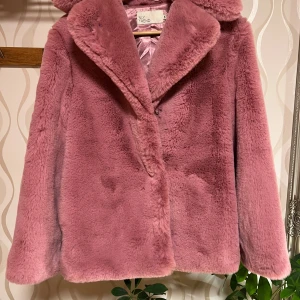 Rosa pälsjacka från NLY Trend - Säljer en fluffig och mjuk rosa pälsjacka från NLY Trend i storlek 34. Jackan har bred krage, knappar framtill och ett glansigt rosa foder. Perfekt för dig som vill ha en statement piece med cozy vibes och snygg siluett.