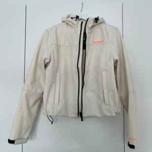 Beige vindjacka från Superdry - Snygg beige vindjacka från Superdry med huva och svarta detaljer. Jackan har dragkedja framtill, snörning i midjan och två fickor. Logotypen syns i neonorange på bröstet och ryggen. Perfekt för blåsiga dagar och sportiga outfits.