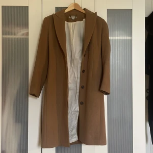 Brun kappa från H&M med bälte, stl 36 - Snygg brun kappa från H&M i storlek 36. Klassisk modell med knappar framtill, bälte i midjan och mjukt innerfoder. Perfekt för höst och vinter när du vill ha en stilren look. Långa ärmar och enkel krage ger en clean vibe.