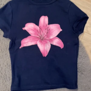 Mörkblå t-shirt med rosa blomma  NEWYORKER - Mörkblå t-shirt från FB Sister i storlek S med en stor rosa blomma tryckt på framsidan. Klassisk rund hals och korta ärmar. Perfekt för dig som vill sticka ut med en färgglad och unik detalj. Gjord i mjuk bomull för skön känsla hela dagen.