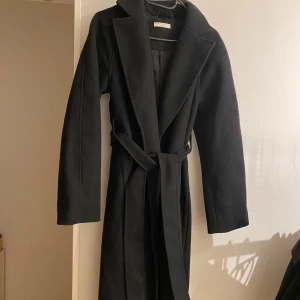 Svart lång kappa - Stilren svart kappa från Gina Tricot med klassisk krage och knytskärp i midjan. Lång modell med slits bak och diskreta fickor i sidorna. Perfekt för dig som gillar en clean och tidlös look. Materialet är mjukt och känns lyxigt. Endast använd några få gånger under förra hösten, finns lite noppror och lite smink fläckar på kragen. Nypris 799
