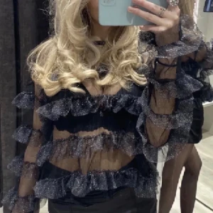 Glittrig svart blus med volanger - Säljer en glittrig svart blus med transparent mesh och flera lager volanger både på ärmar och över kroppen. Blusen har långärmad design och ger en riktigt cool och festlig vibe. Perfekt för dig som vill sticka ut med en unik topp!