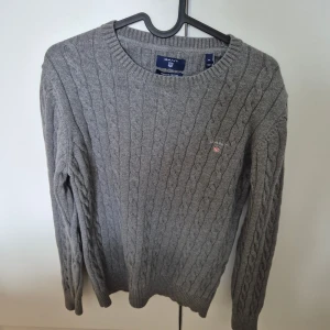 Grå kabelstickad tröja från Gant - Säljer en grå kabelstickad tröja från Gant i premium bomull. Tröjan har rund halsringning, lång ärm och Gant-logga broderad på bröstet. Perfekt för dig som gillar klassisk och stilren look.