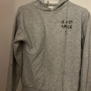 Grå hoodie från Gina Tricot med tryck - Grå hoodie från Gina Tricot med svart texttryck 'Je m'en amour' och en stjärna både fram och bak. Mjuk insida, huva och långa ärmar. Perfekt för dig som gillar en chill och enkel stil.