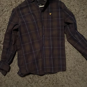 Rutig skjorta från Lyle & Scott - Snygg rutig skjorta från Lyle & Scott i mörkblå och bruna toner med klassisk krage och långa ärmar. Skjortan har knappar framtill och den ikoniska gula loggan broderad på bröstet. Perfekt för dig som gillar stilrena detaljer och mönster.