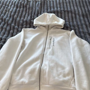 Vit hoodie från Sailracing med dragkedja - Säljer en stilren vit hoodie från Sailracing med hel dragkedja och huva. Tröjan har en bröstficka med dragkedja och diskret logga på bröstet. Perfekt för dig som gillar en clean look och vill ha något bekvämt till vardagen.