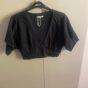 Svart croppad blus från Gina Tricot - Svart croppad blus från Gina Tricot i linne med v-ringning och vida korta ärmar. Snygg detalj med knappar i sidan. Perfekt till höga jeans eller kjol för en trendig look.