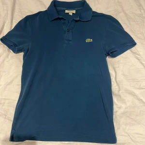 Blå pikétröja från Lacoste slim fit - Snygg blå pikétröja från Lacoste i slim fit-modell. Klassisk krage, korta ärmar och knäppning framtill. Ikoniska krokodilloggan broderad på bröstet. Perfekt för en clean och sportig stil.