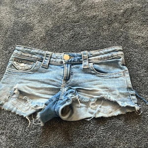 Lågmidjade Ljusblå jeansshorts från Gina Tricot - Säljer ett par lågmidjade ljusblå jeansshorts från Gina Tricot med råa, fransiga kanter och slitna detaljer. Shortsen har klassisk femficksdesign, bälteshällor och knappstängning framtill. Perfekta för varma dagar och en avslappnad look.