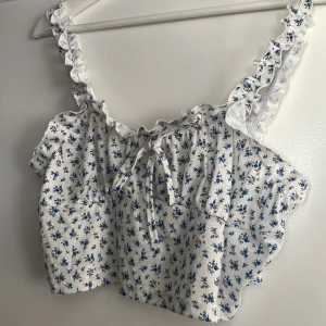 Blommig croppad topp från SHEIN - Supersöt croppad topp från SHEIN med blått blommönster på vit botten. Toppen har volangprydda axelband, knytning framtill och är tillverkad i ett ribbat material. Perfekt för en somrig look med romantiska vibes.