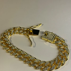 Guldarmband med glittrande stenar - Snyggt chunky armband i guldton med stora länkar täckta av glittrande stenar. Låset är också dekorerat med stenar för extra bling. Perfekt statement piece för dig som vill sticka ut och glänsa lite extra.