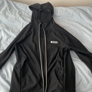 Svart BOSS Matteo Barrettini hoodie - Svart hoodie från BOSS med vit logga på bröstet och hel dragkedja framtill. Snygga vita och beige ränder längs ärmarna ger en sportig vibe. Tröjan har huva och är tillverkad i mjukt polyester-material. Perfekt för chill eller träning.