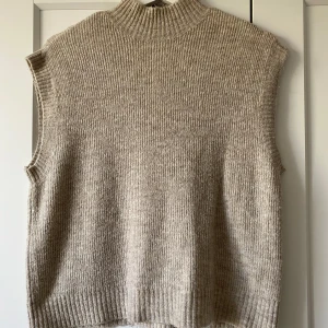 Beige stickad väst från Vero Moda - Stickad väst i beige från Vero Moda med ribbad struktur och hög krage. Modellen är ärmlös och har en rak, loose passform som ger en chill vibe. Perfekt att slänga över en skjorta eller t-shirt för en snygg lager-på-lager-look.