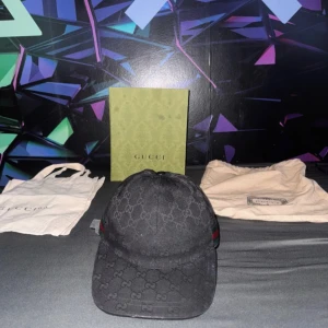 Svart keps från Gucci med GG-mönster - Säljer en svart keps från Gucci med klassiskt GG-monogram över hela kepsen och en diskret röd-grön rand på sidan. Justerbar rem baktill i skinn och insida med Gucci-tag. Kepsen kommer med originalbox och dustbag.