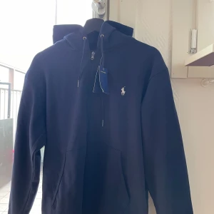 Mörkblå hoodie från Ralph Lauren - Snygg mörkblå hoodie från Ralph Lauren med klassisk logga på bröstet. Tröjan har huva med snörning, halv dragkedja och en stor magficka. 