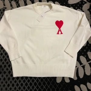 Ami alexandre mattiussi Paris - Vit stickad tröja från Ami Paris med röd hjärtlogga och bokstaven A på bröstet. Tröjan har rund halsringning, ribbade muddar och är långärmad. Perfekt för dig som gillar stilrena plagg med en twist.