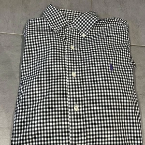 Ralph lauren skjorta - Svartvit rutig skjorta från Ralph Lauren i klassisk passform. Skjortan har lång ärm, knappar framtill och lila broderad logga på bröstet. Tillverkad i mjuk bomull och passar perfekt för en clean och stilren look.