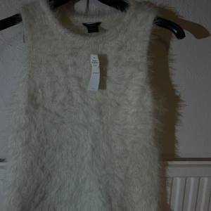 Vit fluffig stickad linnetopp Lindex - Supersöt vit ärmlös stickad linnetopp från Lindex. Toppen har en fluffig, mjuk känsla och är tillverkad i en blandning med återvunnen polyester. Perfekt för lager-på-lager och ger en trendig look med sin unika textur.