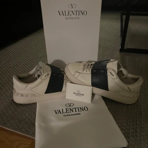 Valentino Garavani sneakers  - Snygga sneakers från Valentino Garavani i vitt med bred svart panel över sidan. Skorna har rund tå, platt sula och klassisk snörning. Perfekta för dig som gillar stilrena och exklusiva sneakers med en modern touch. Kan inte lova att dom är äkta men han jag köpte dom av sa att dom var det!