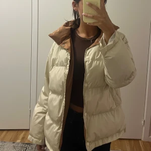 Beige pufferjacka med brun krage - Säljer en beige pufferjacka med brun insida på kragen. Jackan har en oversized passform, långa ärmar och stängs med knappar framtill. 