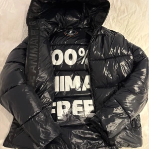 Save The Duck Jacks - Säljer en svart, glansig pufferjacka från Save The Duck med huva och dragkedja. Topp skick förutom texten som hae blivit lite sliten 