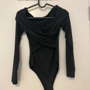 Svart omlott bodysuit med drapering - Svart långärmad bodysuit med snygg omlott-drapering över bysten och axlarna. Djup v-ringning och figurnära passform som framhäver siluetten. Perfekt att styla med jeans eller kjol för en trendig look.