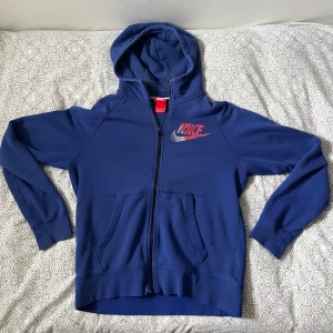 Blå Nike hoodie med dragkedja - Säljer en blå hoodie från Nike med hel dragkedja och huva. Tröjan har Nike-logga i rött och silver på bröstet samt två stora fickor framtill. Perfekt för chill dagar eller när du vill ha en sportig look.