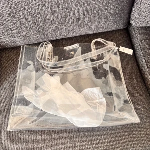 Transparent shoppingväska i syntet - Säljer en stor och trendig transparent shoppingväska i syntetmaterial. Väskan har två långa handtag och stängs med tryckknapp upptill. Perfekt för dig som vill visa upp dina grejer och ha en unik accessoar till vardagslooken.