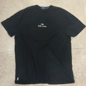 Svart t-shirt från Polo Ralph Lauren - Svart t-shirt från Polo Ralph Lauren med klassisk passform. T-shirten har vit broderad logga på bröstet och liten logga nertill. Tillverkad i mjuk bomull och har rund hals samt korta ärmar.