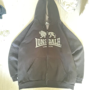 Svart hoodie från Lonsdale med tryck - Svart hoodie från Lonsdale London med stort vitt logotryck på bröstet och två ränder längs ärmarna. Tröjan har huva med snörning, dragkedja framtill och känguruficka. Perfekt för en sportig och avslappnad stil.