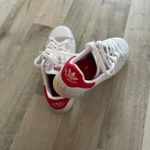 Adidas Stan Smith – vita sneakers med röda detaljer! - Tidlösa Stan Smith i vitt skinn med röda detaljer. Perforerade sidor, snörning och platt sula för en clean, sportig look. Perfekta till allt!