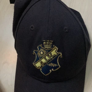 AIK keps från New Era 9FORTY - Svart AIK keps från New Era 9FORTY med broderad AIK-logga i guld och blått framtill. Justerbar rem baktill för perfekt passform. Klassisk böjd skärm och broderad New Era-logga på sidan. Perfekt för dig som vill visa ditt AIK-stöd.