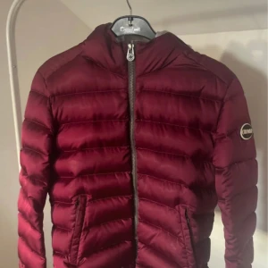 Vinröd dunjacka från Colmar - Stilren och lätt dunjacka från Colmar i en elegant vinröd nyans. Klassisk  design som håller dig varm utan att kännas tung.  🎨 Färg: Vinröd 📏 Storlek: 42 (motsvarar Small) 📐 Mått:fråga om önskas  ♻️ Skick:8/10 🧵 Material:  100% nylon, fodrad med dun och fjäder.