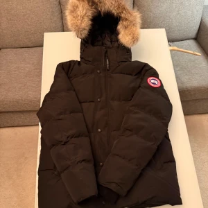 Svart dunjacka från Canada Goose L - Säljer en svart dunjacka från Canada Goose med stor huva och snygg pälsdetalj. Jackan har klassisk patch på ärmen, dragkedja och knappar framtill. Perfekt för kalla vinterdagar och riktigt varm. Stilsäker och tidlös modell.